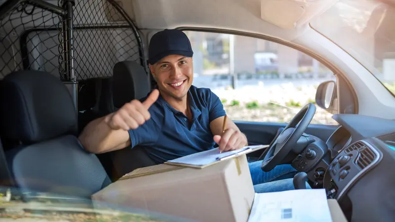 Zusteller im Lieferwagen, ein Paket auf dem Beifahrersitz, hebt lächelnd den Daumen