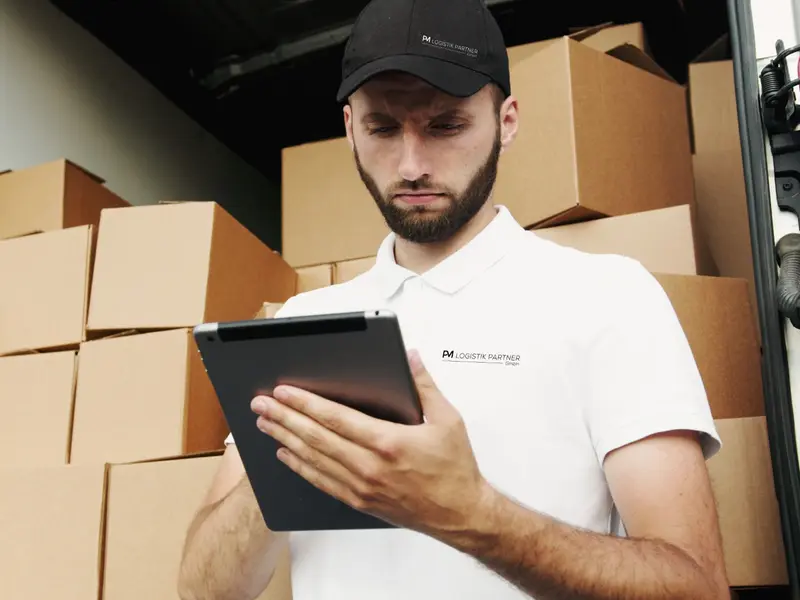 Zusteller in Cap und Polo mit PM-Logo prüft vor dem beladenen Lieferwagen die Tour auf dem Tablet